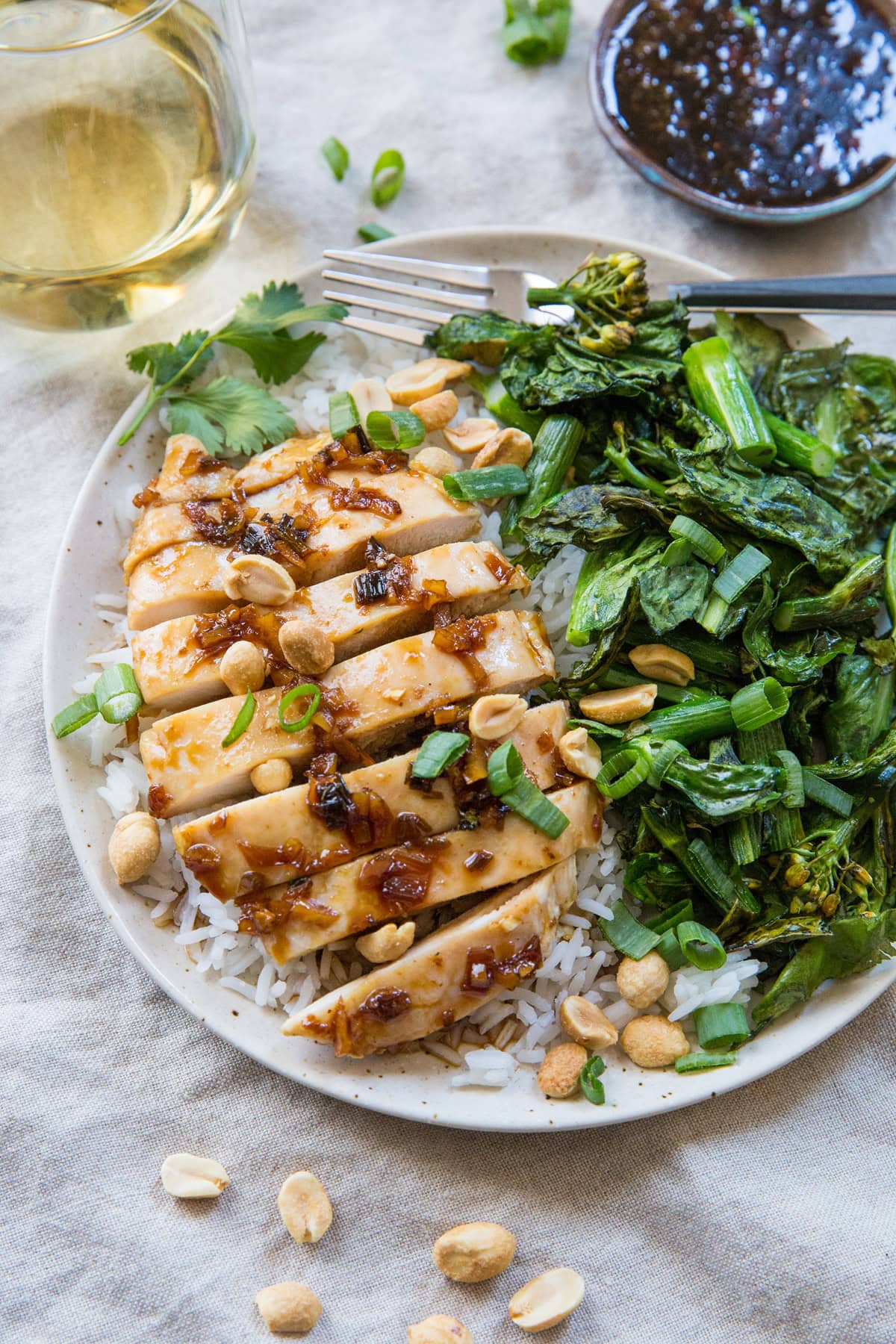 lemongrass chicken on a plate with white rice, broccoli rabe, sauce in a bowl in the background and a glass of wine in the background.