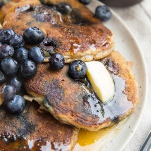 Almond flour pancakes on a plate with fresh blueberries, melted butter, and honey. A fork to the side, ready to serve.