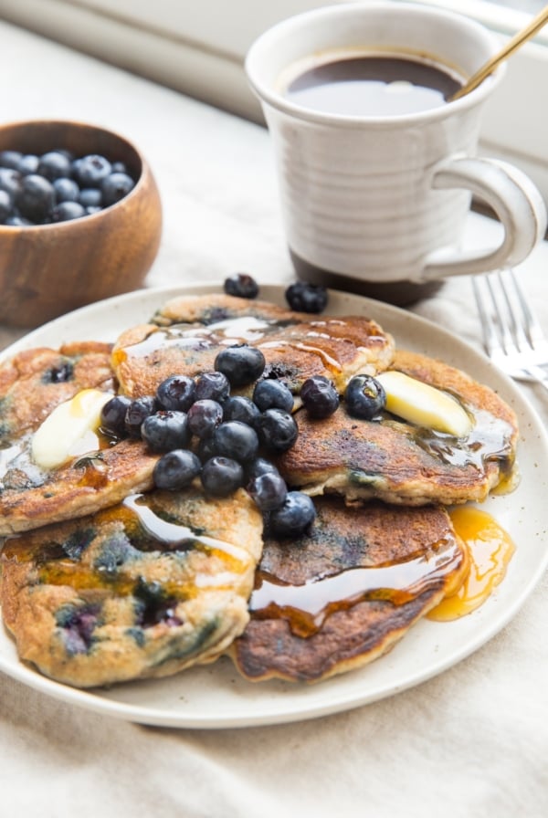Plate of five blueberry pancakes with melted butter, honey drizzled on top and fresh berries. A mug of coffee and a bowl of blueberries in the background.