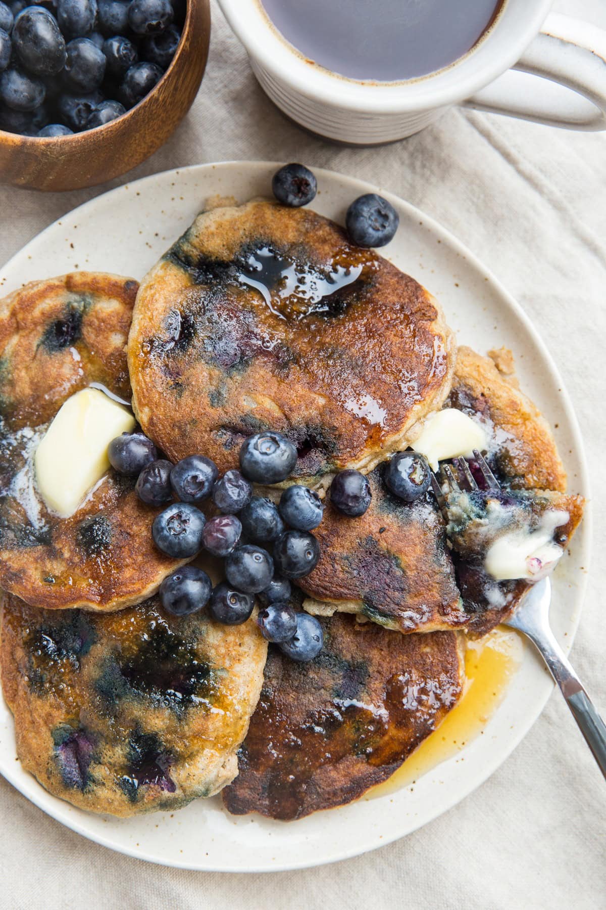 Plate of five blueberry pancakes with a fork taking a bite out of one of them. A mug of coffee and fresh blueberries to the side.