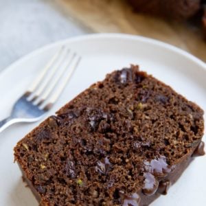 Slice of chocolate zucchini bread on a white plate with the rest of the loaf sliced in the background.
