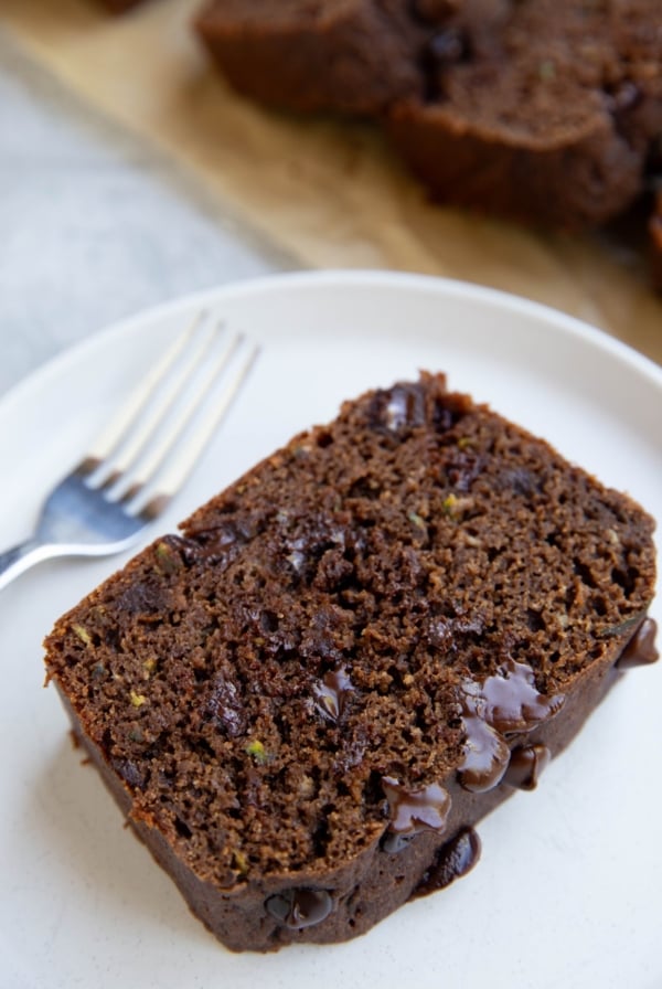 Slice of chocolate zucchini bread on a white plate with the rest of the loaf sliced in the background.