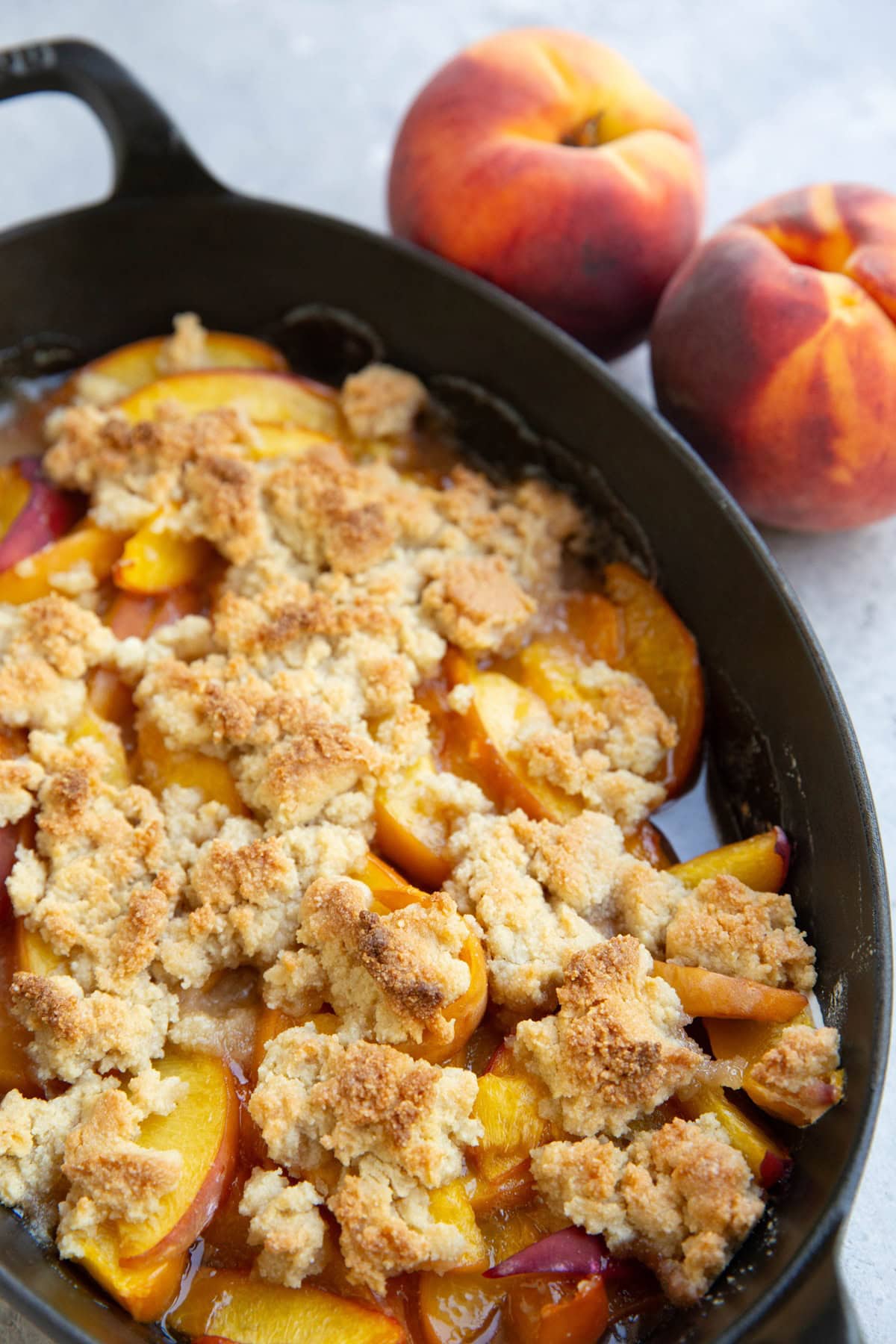 Baking dish of peach crisp with fresh peaches in the background, fresh out of the oven and ready to serve.