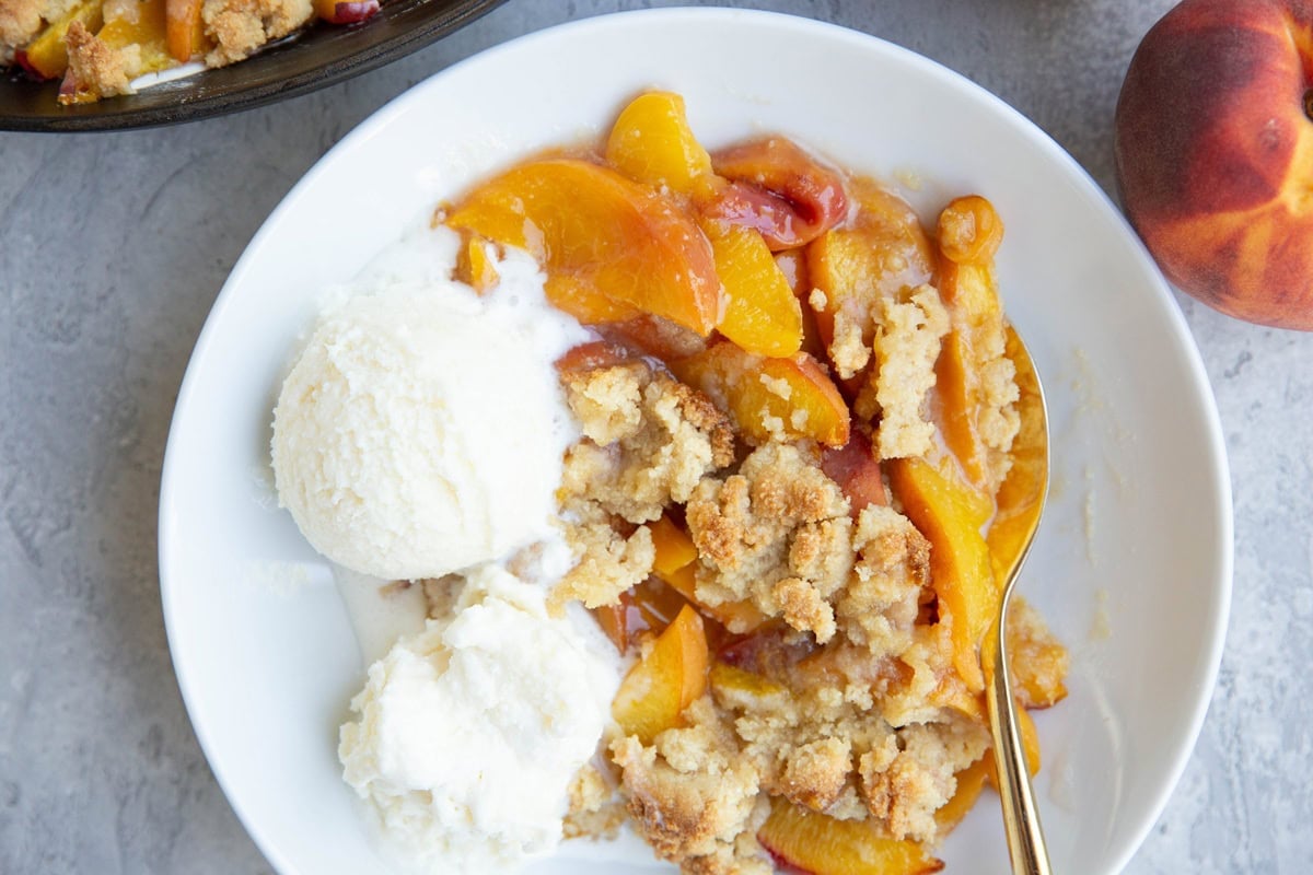 White bowl of peach crisp with scoops of vanilla ice cream to the side. a fresh peach to the side and a gold spoon with the crumble.