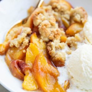 Peach crisp in a white bowl with a scoop of melty vanilla ice cream and a golden spoon to the side, ready to eat.