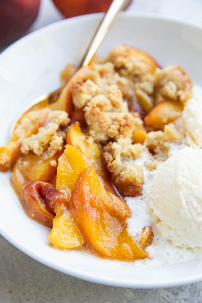 Peach crisp in a white bowl with a scoop of melty vanilla ice cream and a golden spoon to the side, ready to eat.