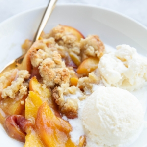 Peach crisp in a white bowl with scoops of vanilla ice cream to the side. A golden spoon next to the fruit crisp, ready to eat.