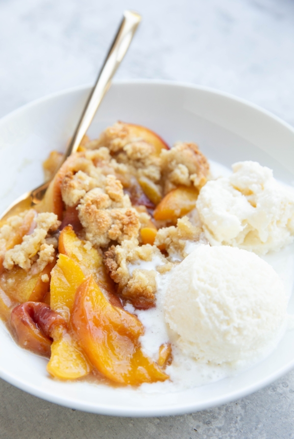 Peach crisp in a white bowl with scoops of vanilla ice cream to the side. A golden spoon next to the fruit crisp, ready to eat.