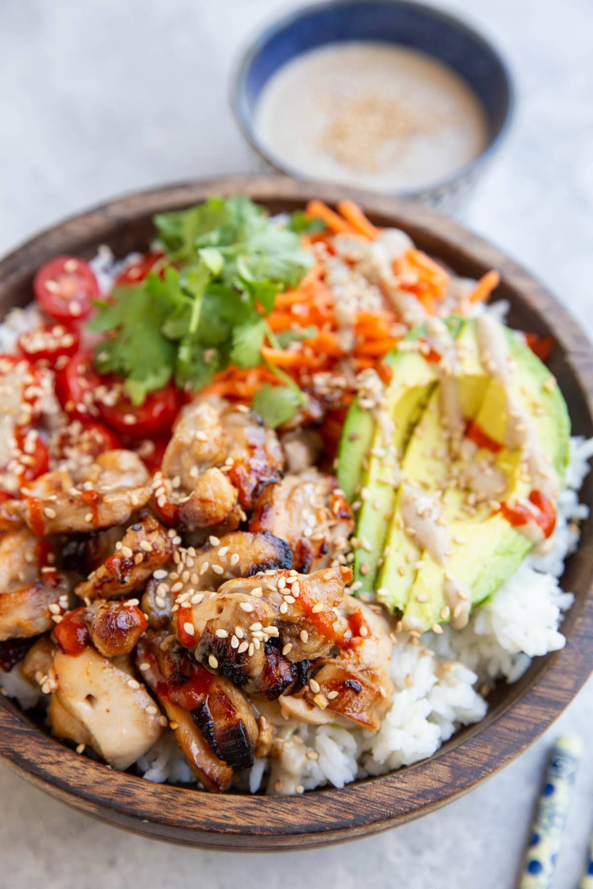 Wooden bowl full of white rice and chicken satay with avocado, grated carrots, tomatoes, and a drizzle of peanut sauce.
