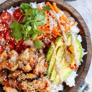 Big wooden bowl full of grilled chicken, rice, avocado, carrots, cherry tomatoes, drizzled with peanut sauce and sriracha. A bowl of peanut sauce and chopsticks to the side.