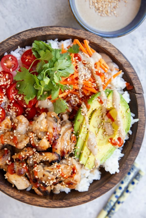 Big wooden bowl full of grilled chicken, rice, avocado, carrots, cherry tomatoes, drizzled with peanut sauce and sriracha. A bowl of peanut sauce and chopsticks to the side.
