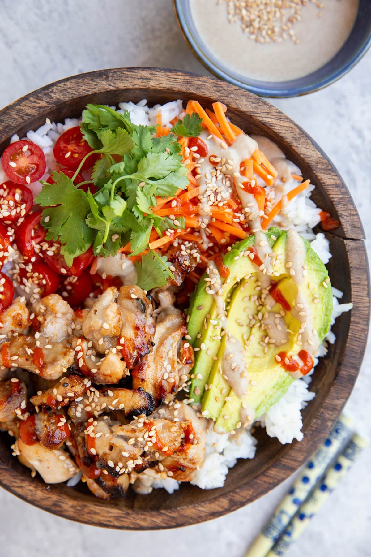 Big wooden bowl full of grilled chicken, rice, avocado, carrots, cherry tomatoes, drizzled with peanut sauce and sriracha. A bowl of peanut sauce and chopsticks to the side.
