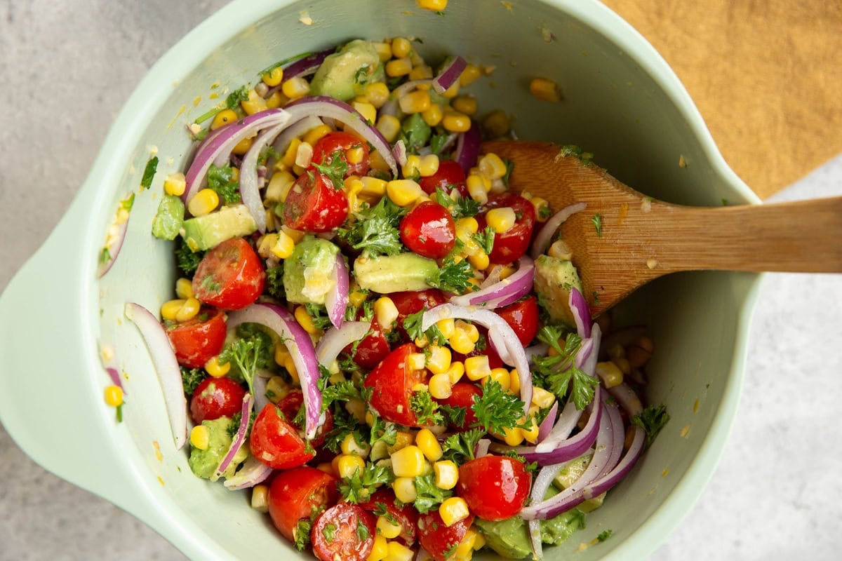 Mixing bowl with tomato and corn salad mixed together with a wooden spoon.