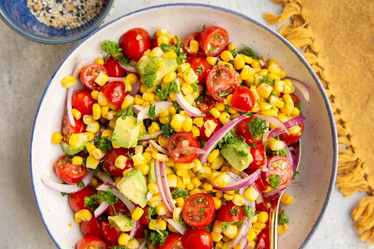 Blue-rimmed white bowl full of corn and tomato salad with a golden napkin to the side.