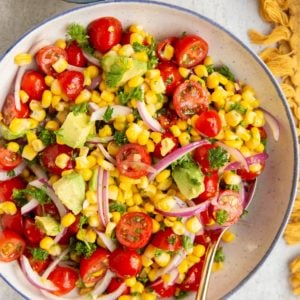 Large bowl full of corn and tomato salad with a golden spoon to the side, a napkin, and a bowl of seasonings.