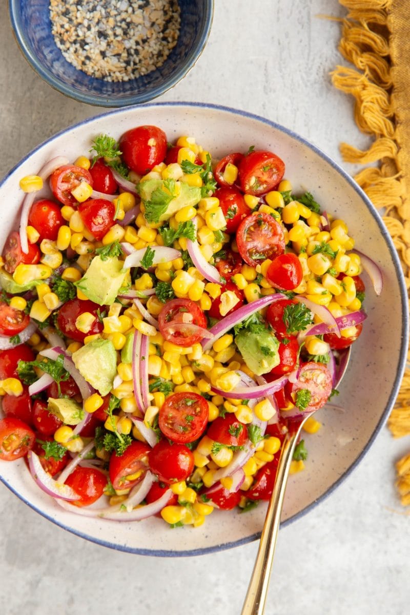 Large bowl full of corn and tomato salad with a golden spoon to the side, a napkin, and a bowl of seasonings.