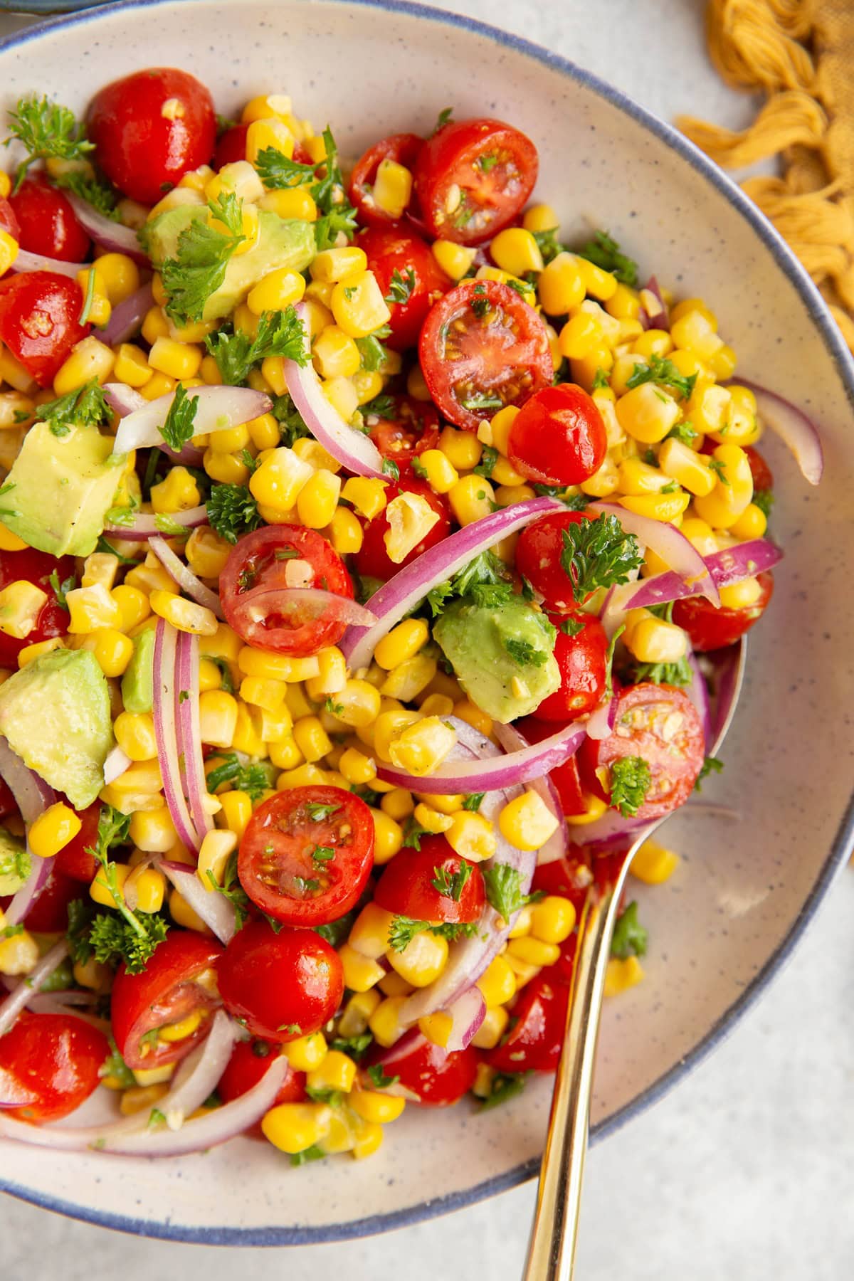 Large bowl full of tomato and corn salad with red onions, avocado, parsley, and more. A golden spoon to the side, ready to serve the side dish.