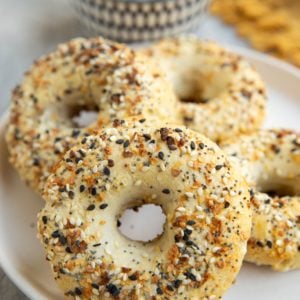 Four homemade bagels on a plate with a bowl of everything bagel seasoning in the background.