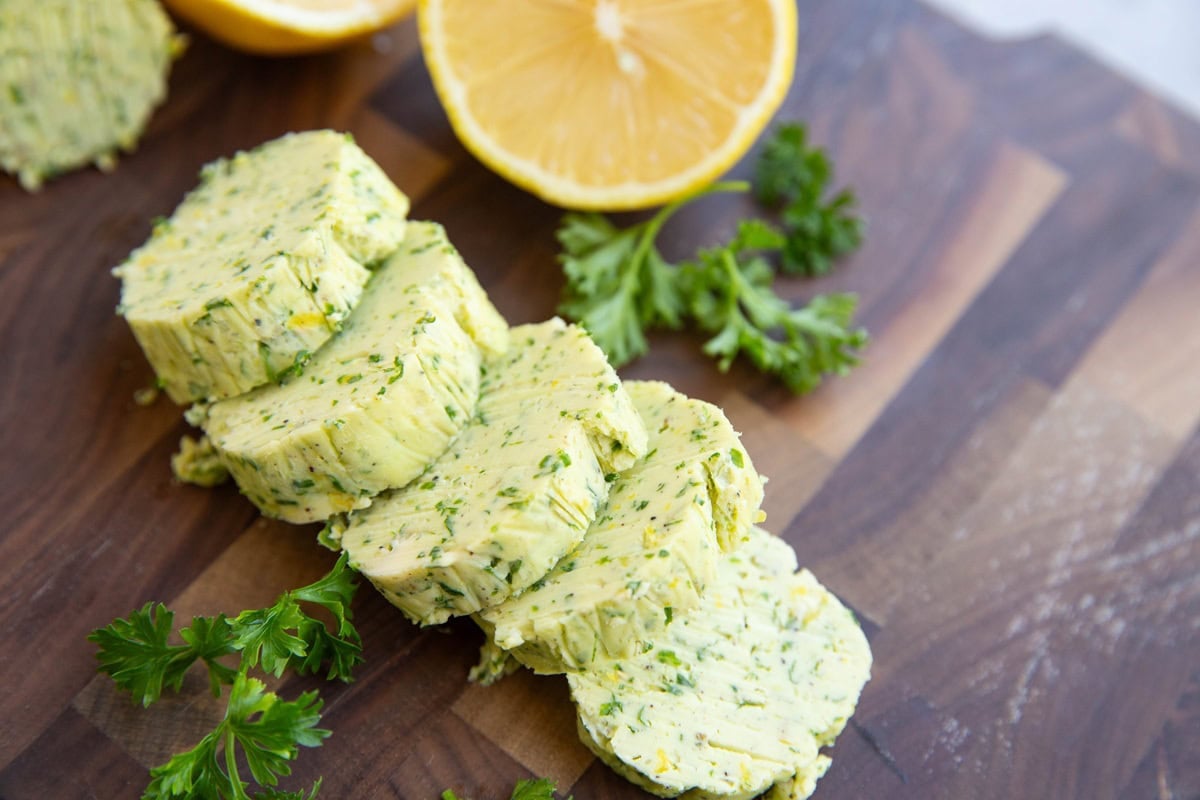 Compound butter on a wooden cutting board cut into slices with fresh herbs and fresh lemon to the side.