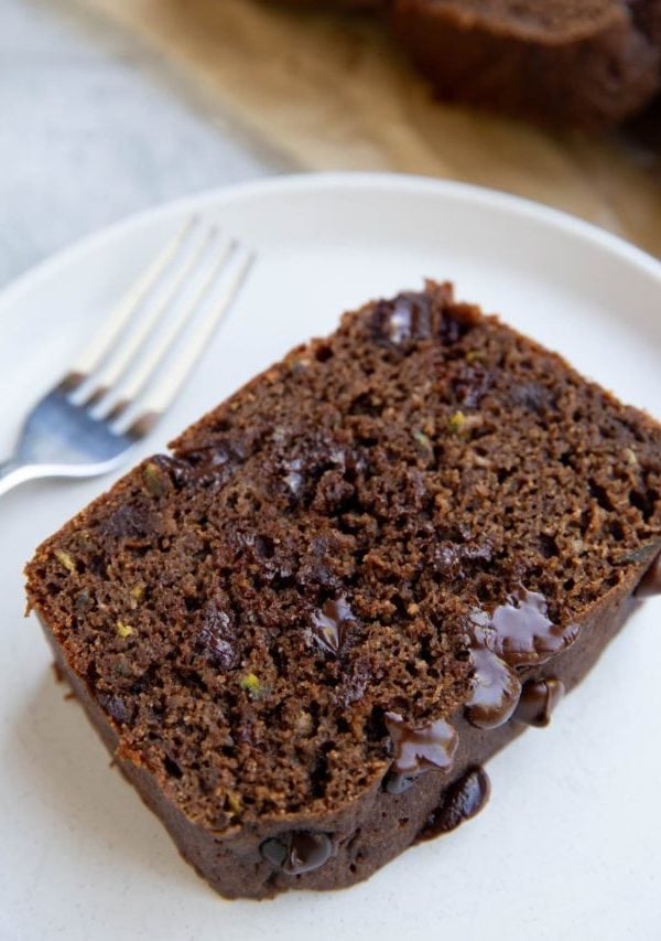 Slice of chocolate zucchini bread on a white plate with the rest of the loaf sliced in the background.