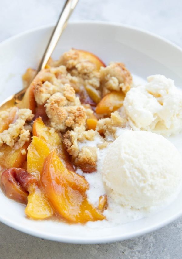 Peach crisp in a white bowl with scoops of vanilla ice cream to the side. A golden spoon next to the fruit crisp, ready to eat.