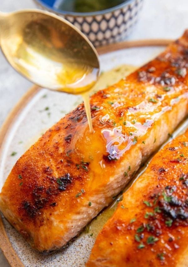Two salmon filets on a plate with a golden spoon drizzling honey butter over the salmon.