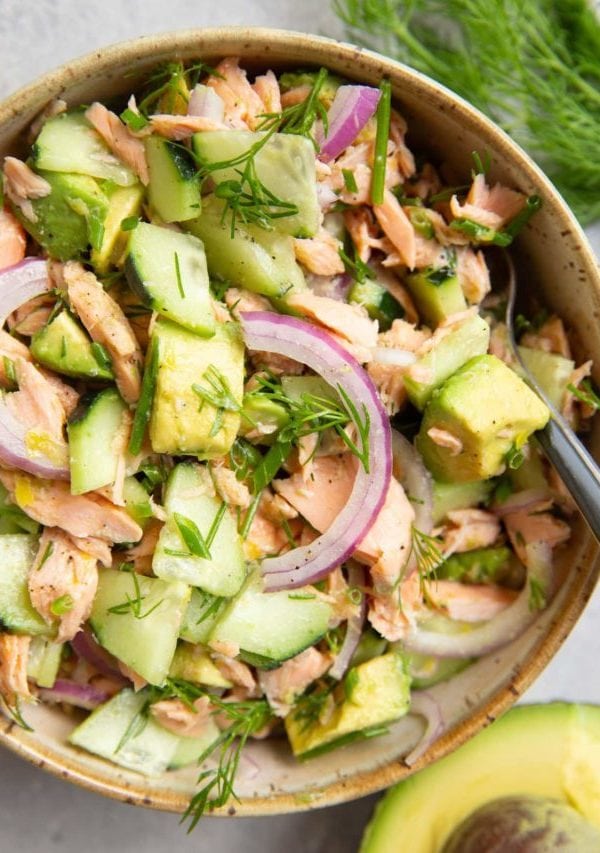 Bowl of cucumber dill avocado salmon salad with a fork ready to serve, half an avocado to the side and fresh dill to the side.