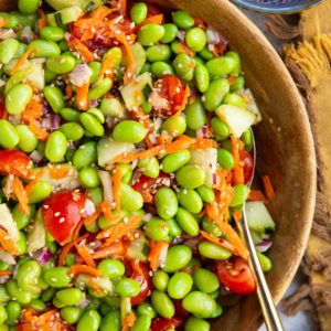 Edamame salad in a wooden bowl with a golden spoon and a blue bowl of sesame seeds to the side.