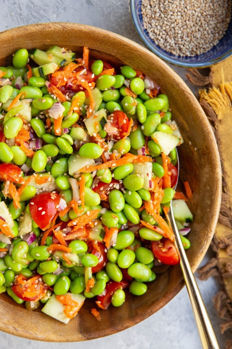 Edamame salad in a wooden bowl with a golden spoon and a blue bowl of sesame seeds to the side.