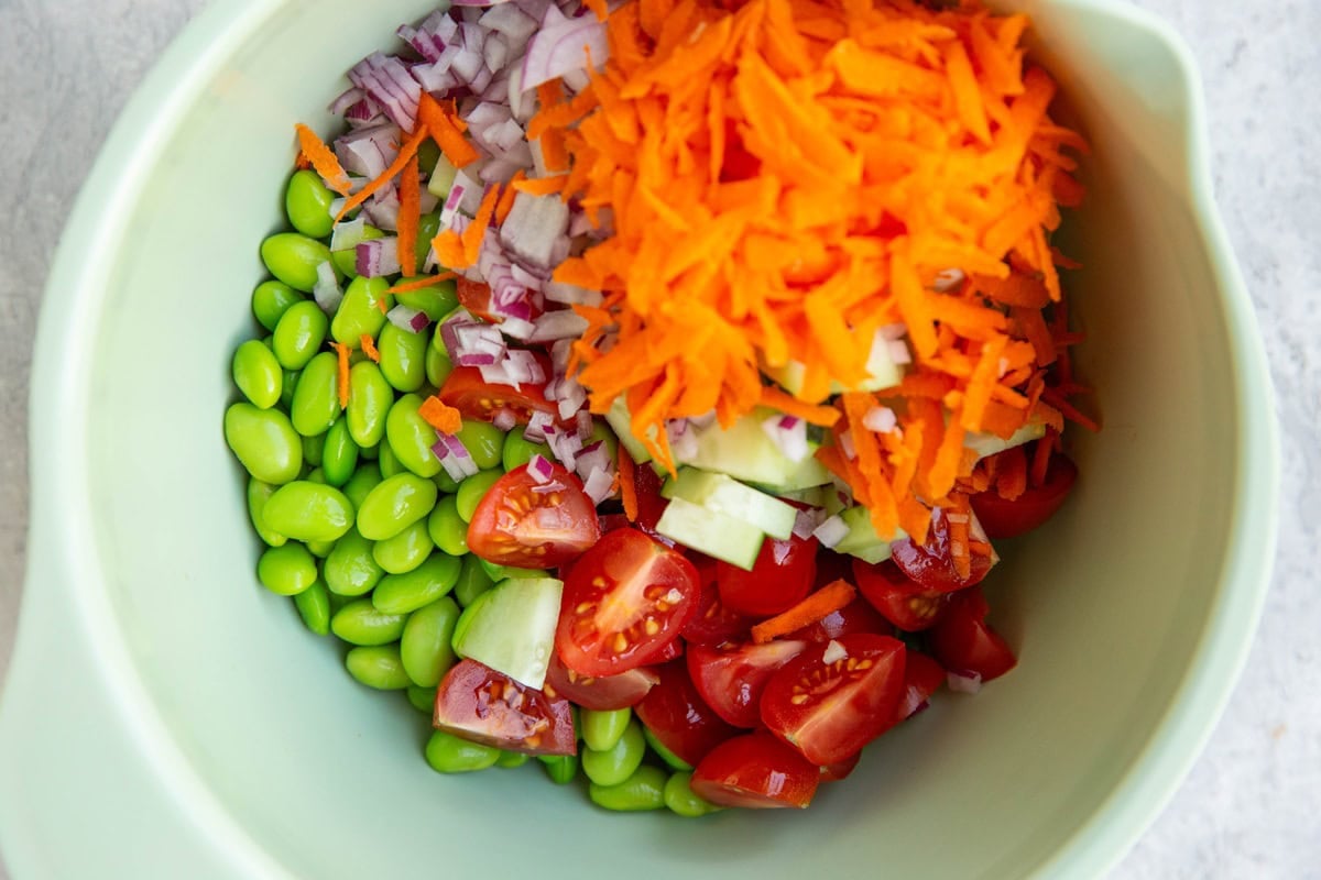Mixing bowl full of edamame, tomatoes, cucumber, red onion, and carrots.