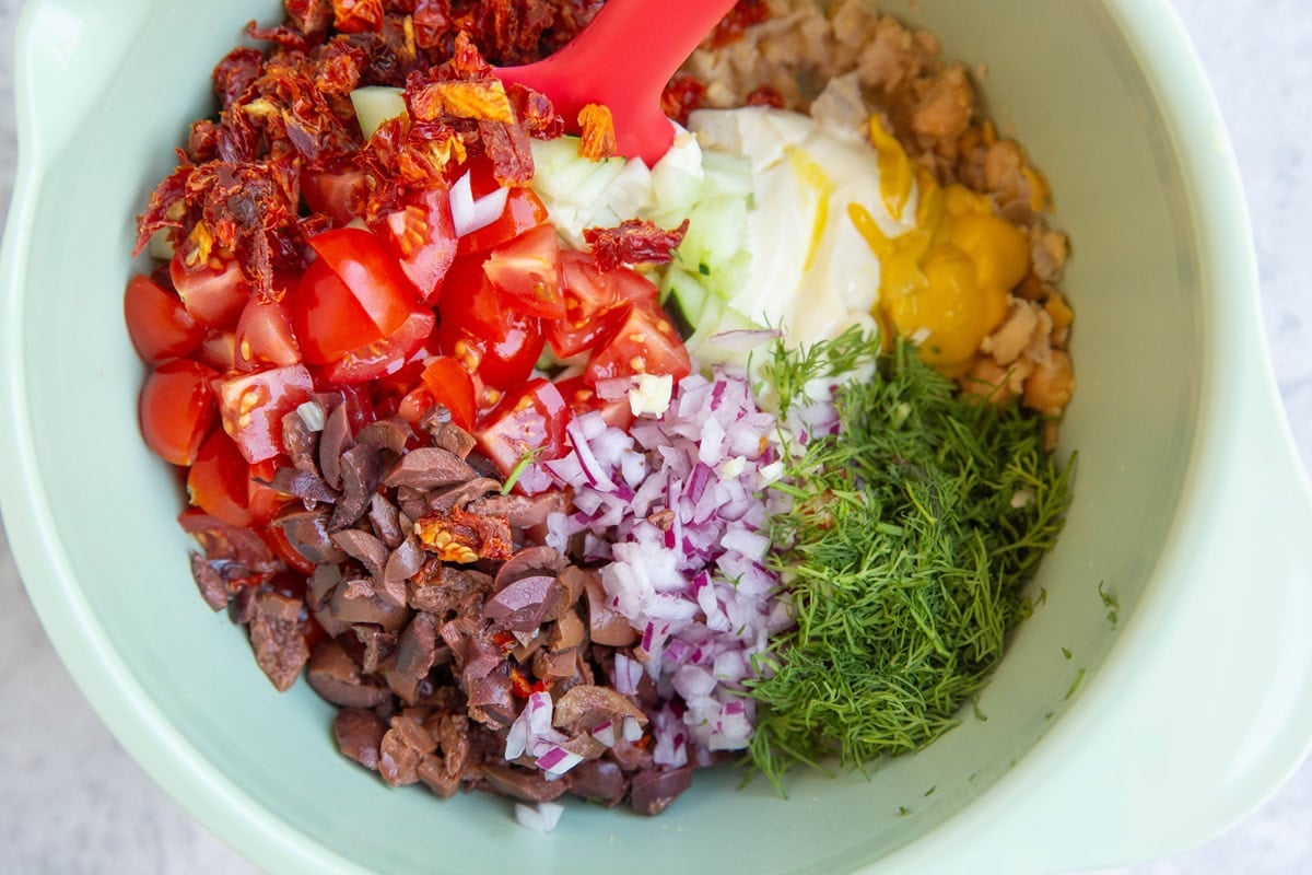 Mashed chickpeas, mayonnaise, cucumber, sun-dried tomatoes, kalamata olives, dill, mustard, and cherry tomatoes in a mixing bowl to be mixed