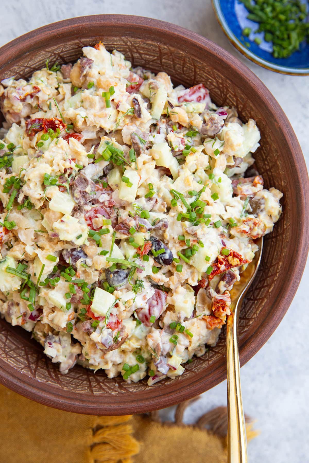 Bowl full of chickpea tuna salad with a golden spoon to the side and a golden napkin with a bowl of chopped chives.