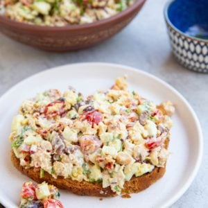 Bread with chickpea tuna salad on top on a white plate and the rest of the salad in a bowl in the background. A golden napkin to the side.