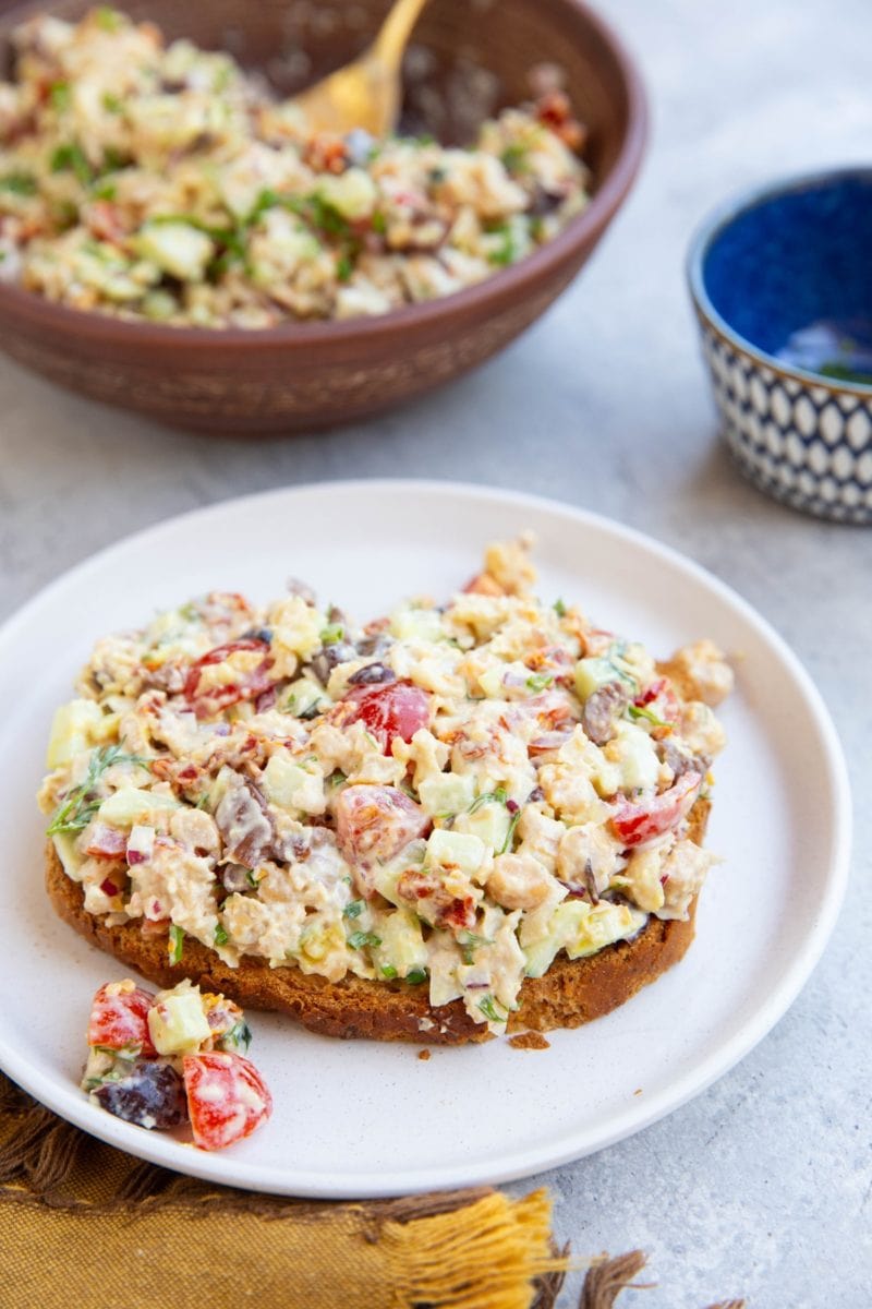 Bread with chickpea tuna salad on top on a white plate and the rest of the salad in a bowl in the background. A golden napkin to the side.