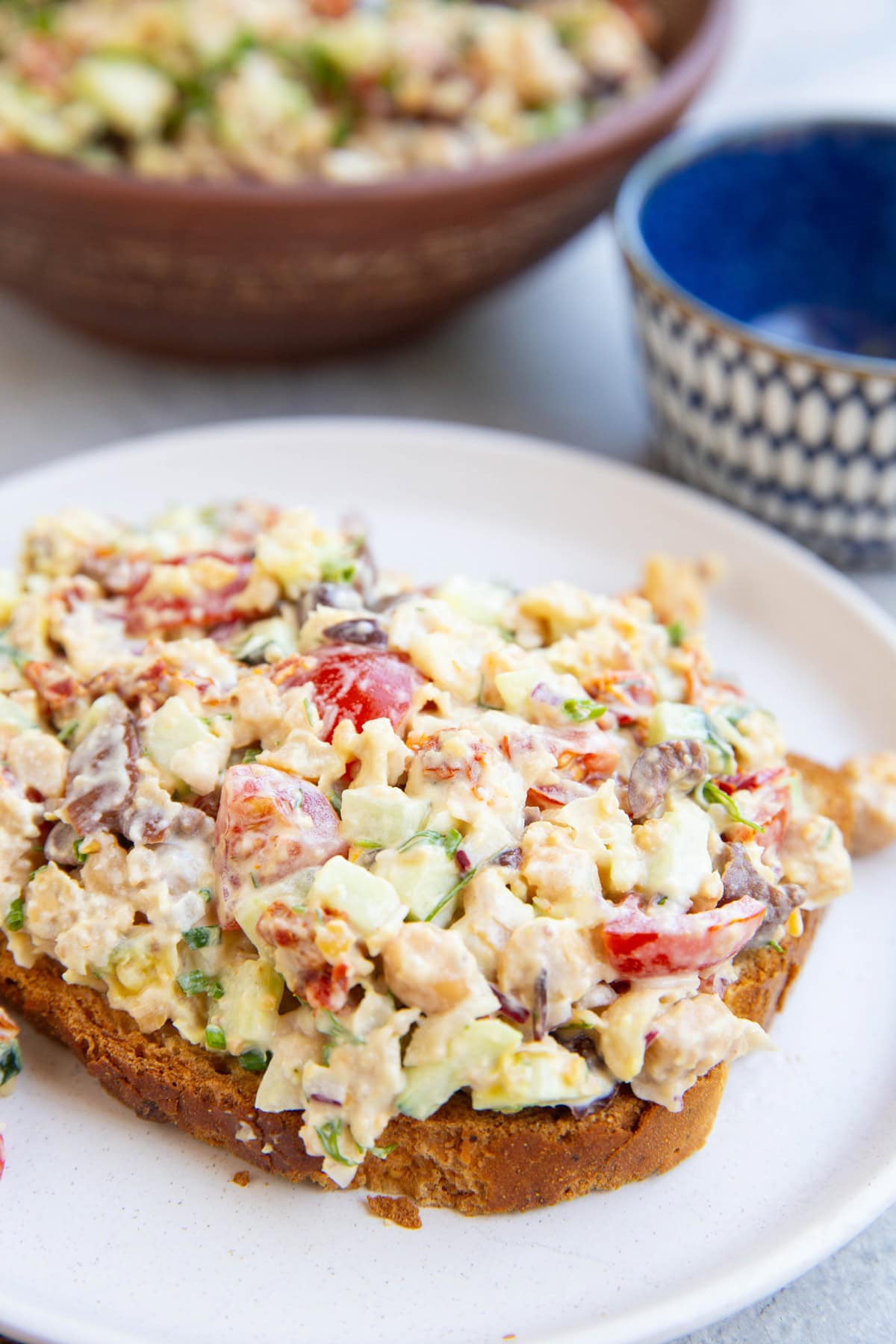 Bread with Greek chickpea tuna salad on top, close up so you can see the individual ingredients. The bowl with the rest of the salad in the background.
