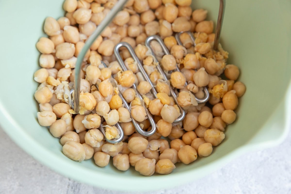 Mixing bowl full of chickpeas being mashed by a potato masher to make salad.
