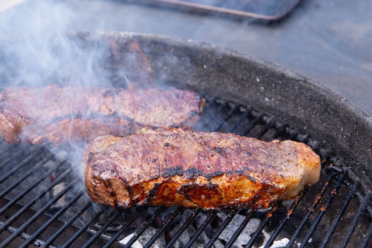 Steaks grilling on a charcoal grill with smoke and flames billowing up.