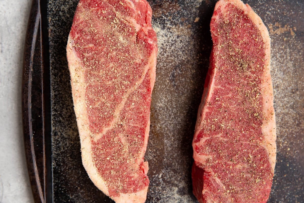 New York strip steaks on a baking sheet, sprinkled with salt, pepper, and garlic powder. Ready to be grilled.