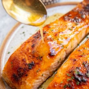 Two salmon filets on a plate with a golden spoon drizzling honey butter over the salmon.