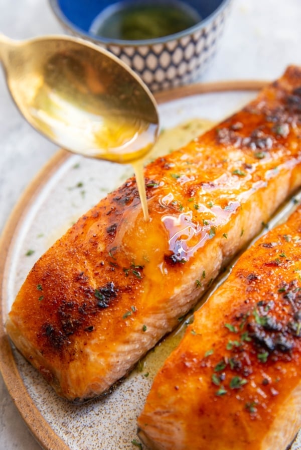 Two salmon filets on a plate with a golden spoon drizzling honey butter over the salmon.