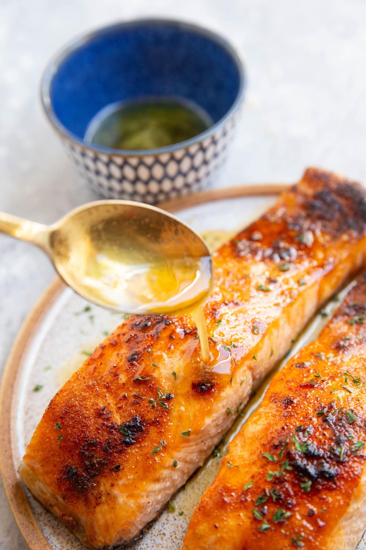 Drizzling hot honey and butter over two cooked salmon filets. A small bowl of hot honey in the background.