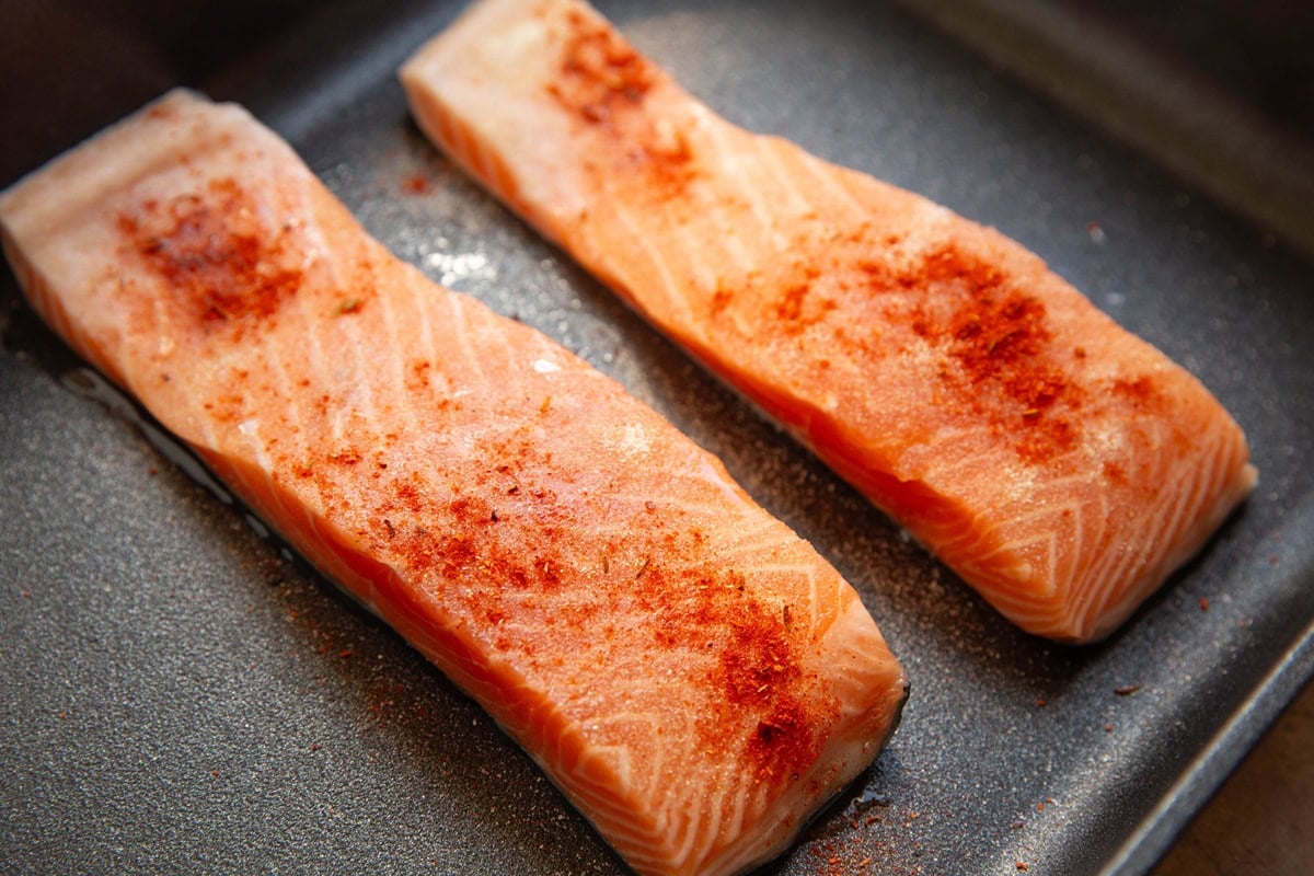 Two raw salmon filets sprinkled with seasonings in a baking dish.