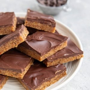 White plate of chocolate chip cookie dough protein bars with a metal measuring cup full of chocolate chips in the background. Bars are ready to eat.