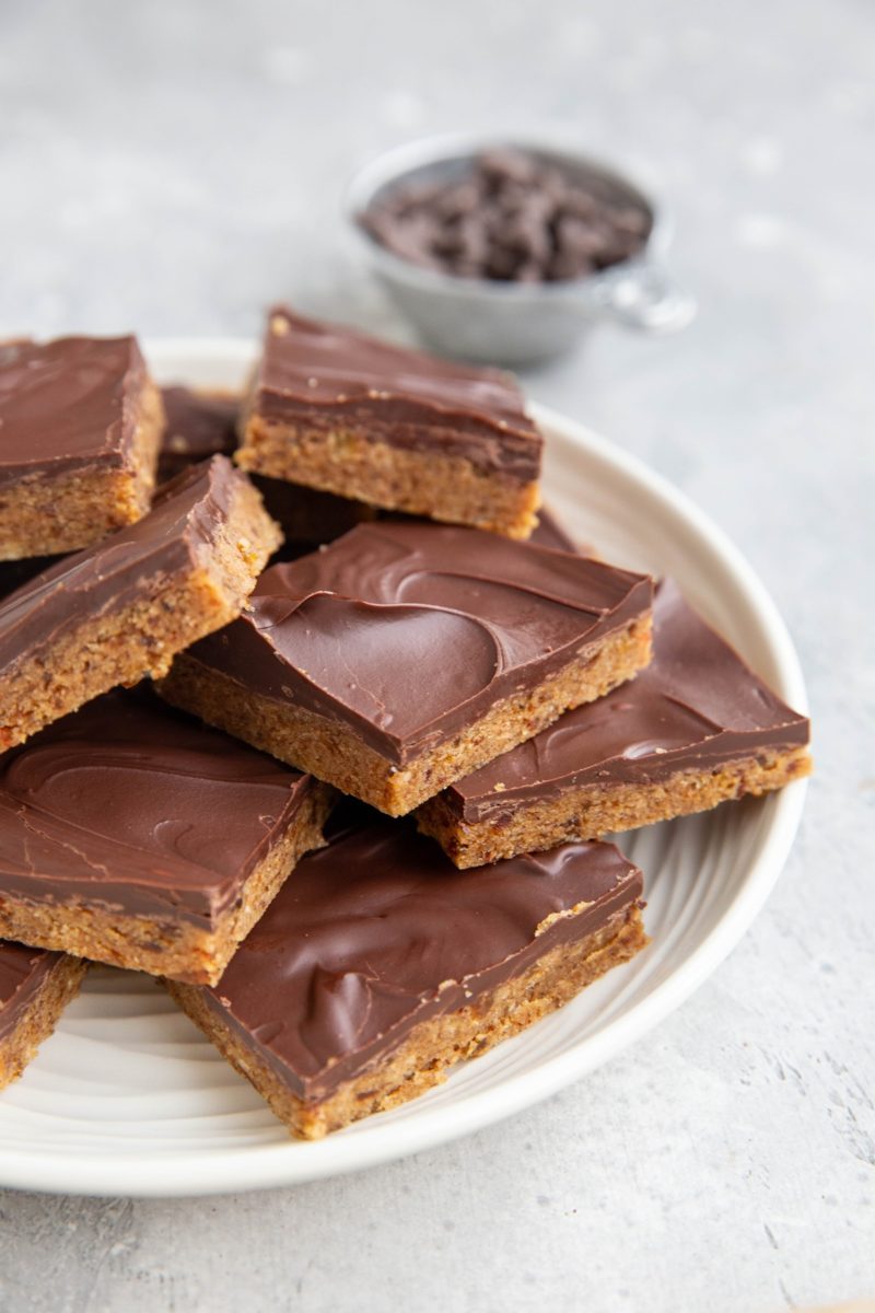 White plate of chocolate chip cookie dough protein bars with a metal measuring cup full of chocolate chips in the background. Bars are ready to eat.