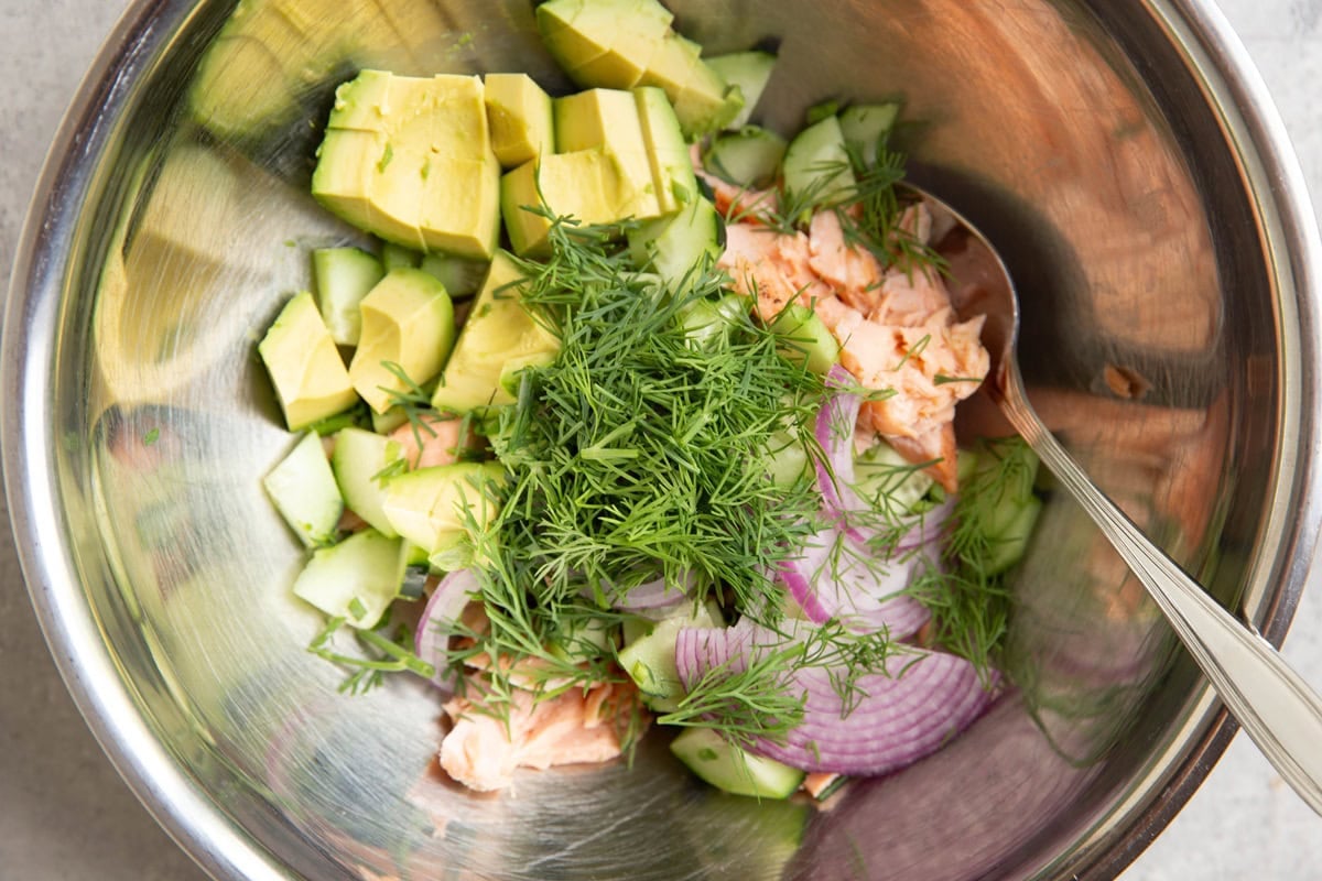 Large stainless steel mixing bowl full of cooked salmon, dill, avocado, cucumbers, and red onion, ready to be mixed together.
