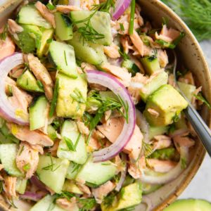 Bowl of cucumber dill avocado salmon salad with a fork ready to serve, half an avocado to the side and fresh dill to the side.