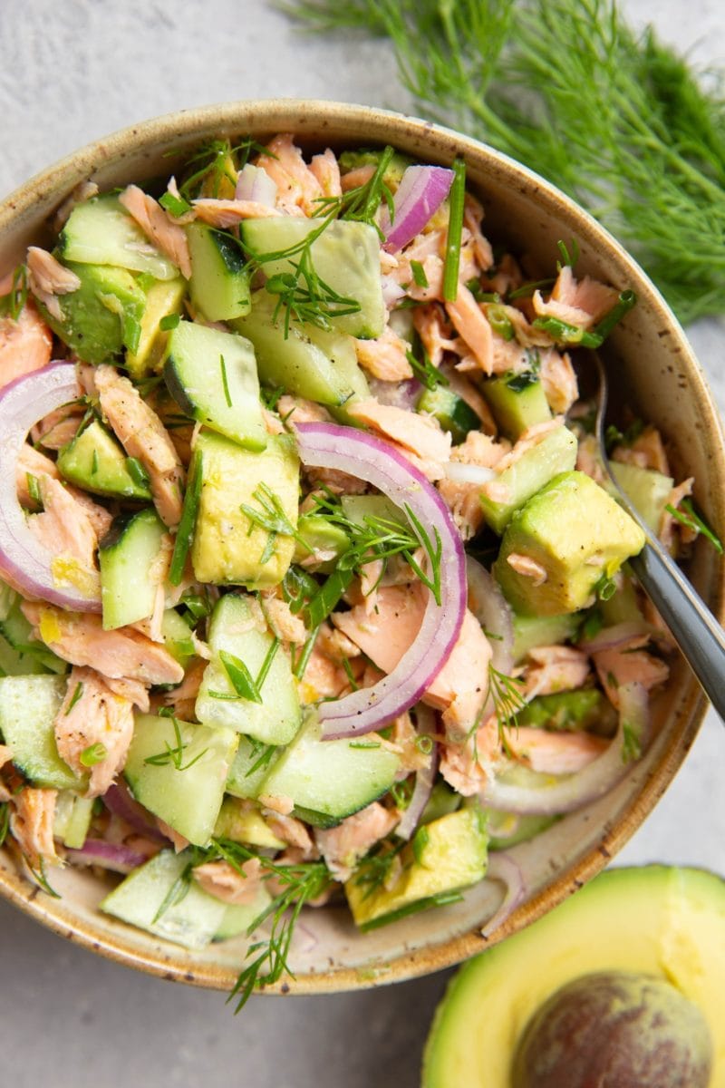 Bowl of cucumber dill avocado salmon salad with a fork ready to serve, half an avocado to the side and fresh dill to the side.