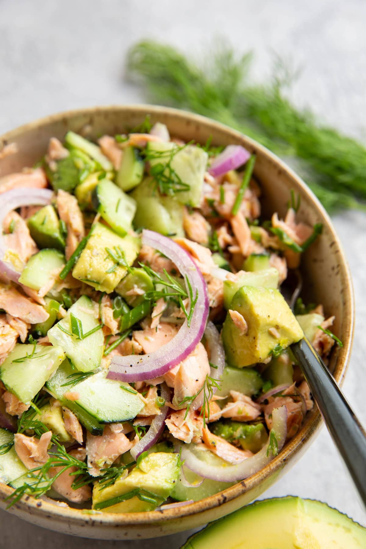 Bowl full of cucumber dill salmon salad with red onion, lemon and a fork to the side. Fresh dill and half of an avocado to the side.