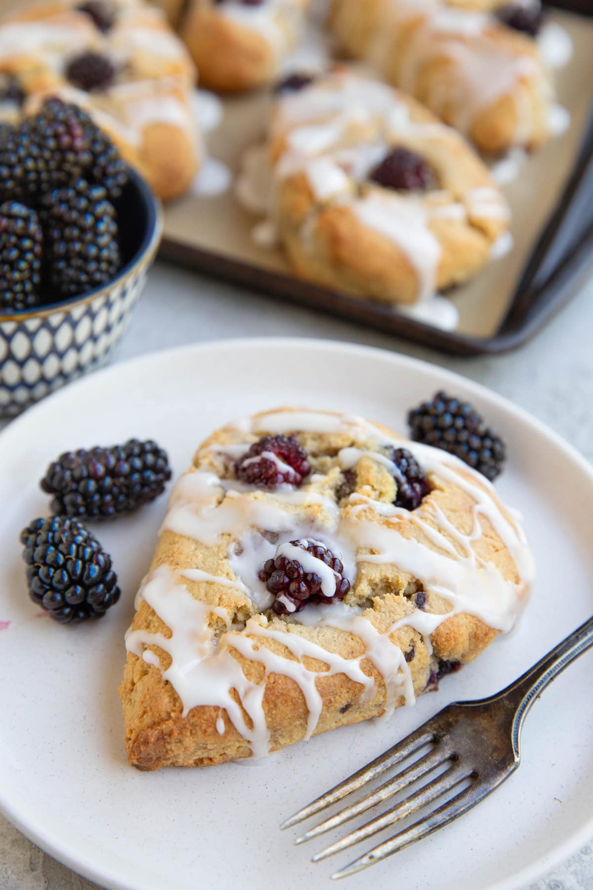 Blackberry scone on a plate with fresh blackberries all around and a baking sheet of scones in the background.