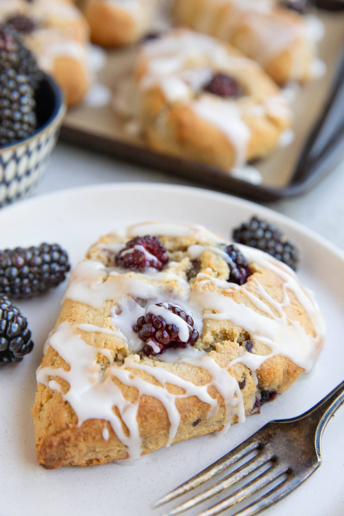 Blackberry almond flour scone on a white plate with fresh blackberries to the side and a fork. The rest of the scones are in the background on a baking sheet and a bowl of fresh blackberries next to it.
